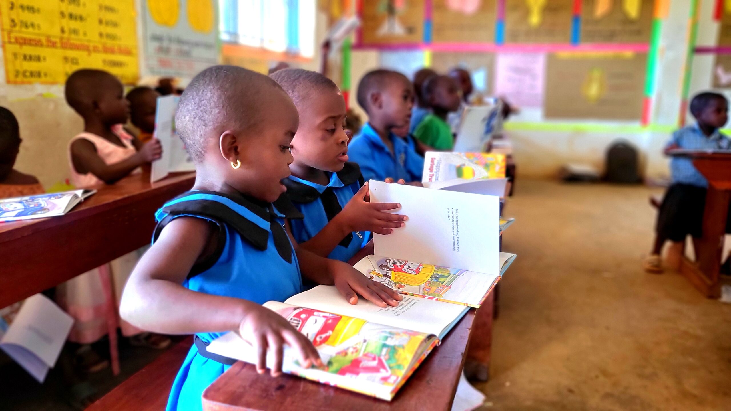 Two young girls in blue FFYEC uniforms read together with focused expressions, sharing a page