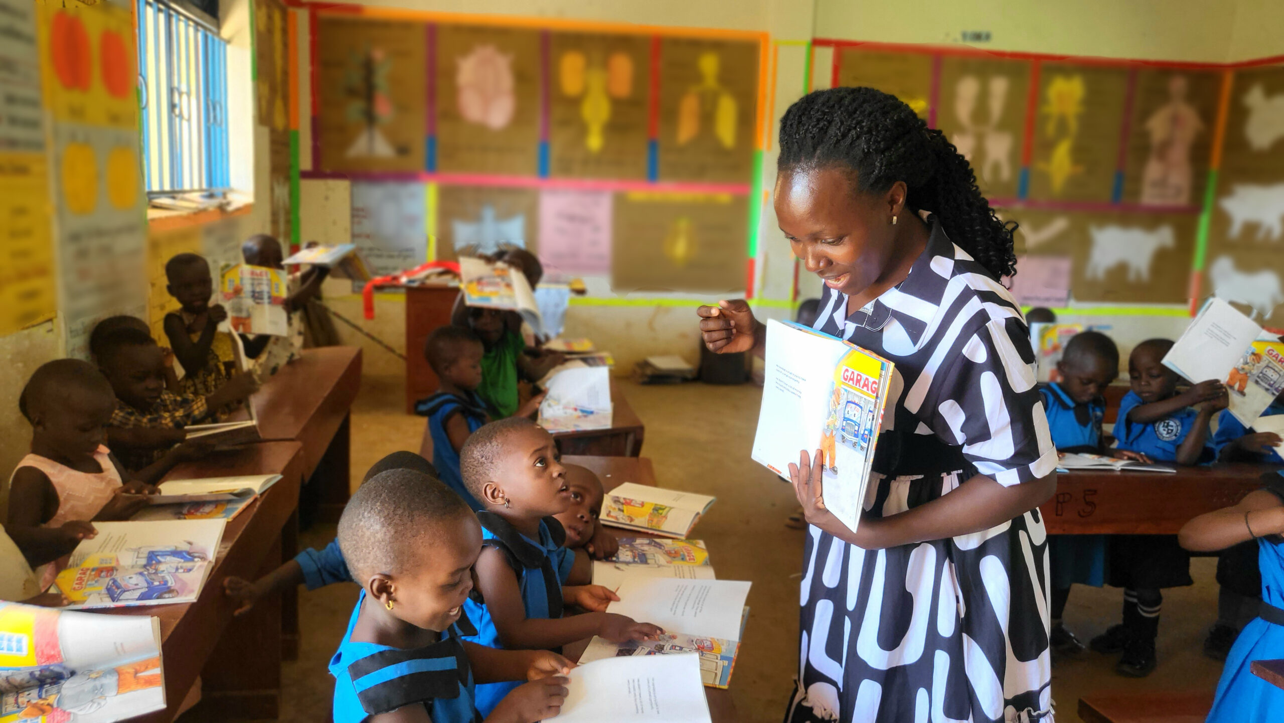 Teacher walks the classroom rows smiling, distributing books to eager learners